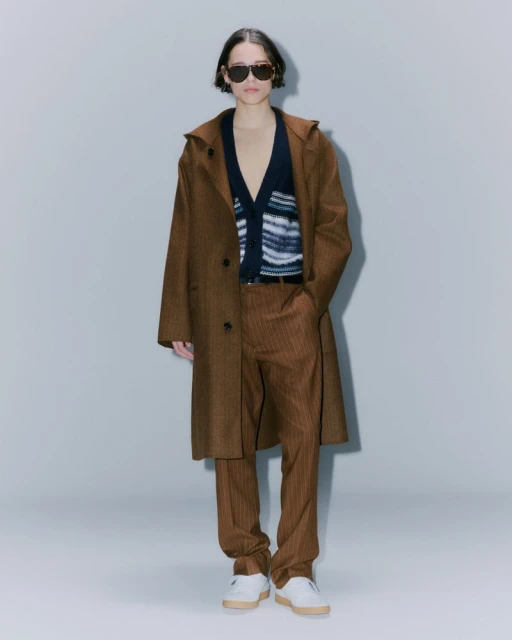 Fashionable person in a brown coat, pinstripe pants, sunglasses, and white sneakers in a full shot against a light background.