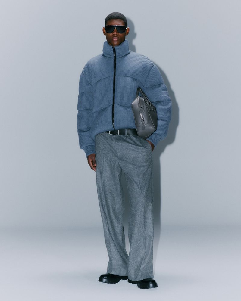 Full-length shot of an adult male wearing a blue puffer jacket, gray pants, sunglasses, and black boots, against a solid gray background.