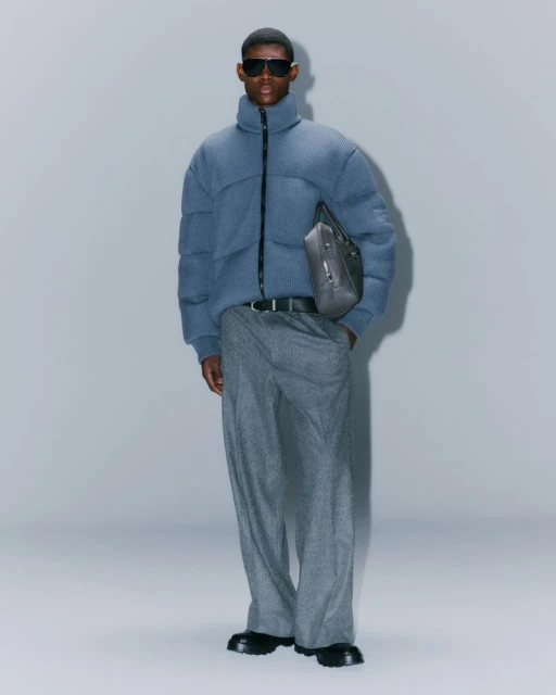 Full-length shot of an adult male wearing a blue puffer jacket, gray pants, sunglasses, and black boots, against a solid gray background.
