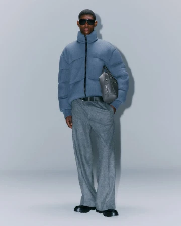 Full-length shot of an adult male wearing a blue puffer jacket, gray pants, sunglasses, and black boots, against a solid gray background.