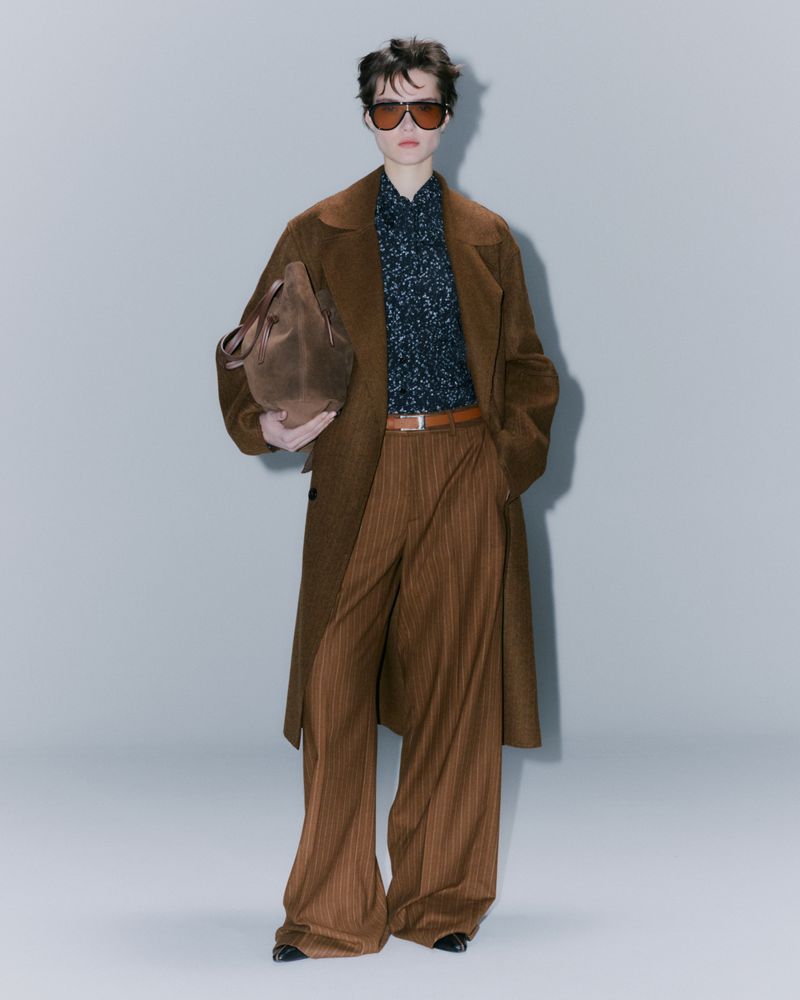 Fashionable adult posing in a long brown coat, patterned shirt, pinstripe trousers, sunglasses and carrying a brown suede tote bag.