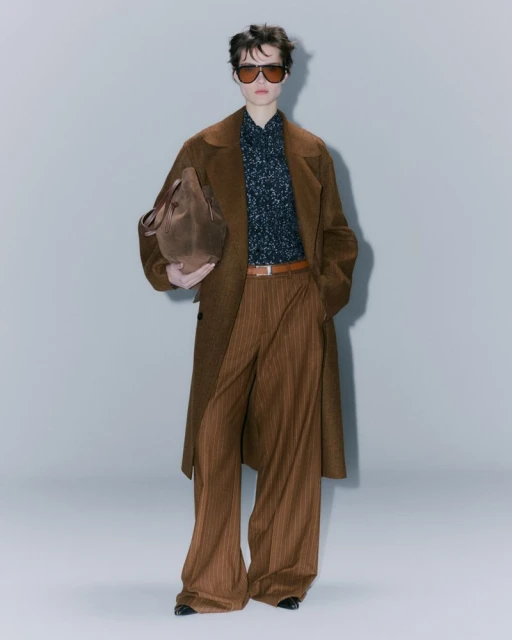 Fashionable adult posing in a long brown coat, patterned shirt, pinstripe trousers, sunglasses and carrying a brown suede tote bag.