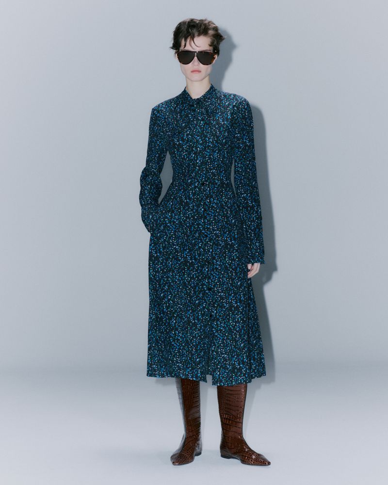 Full-length shot of a stylish woman in a patterned midi dress and brown leather boots with sunglasses against a neutral backdrop.