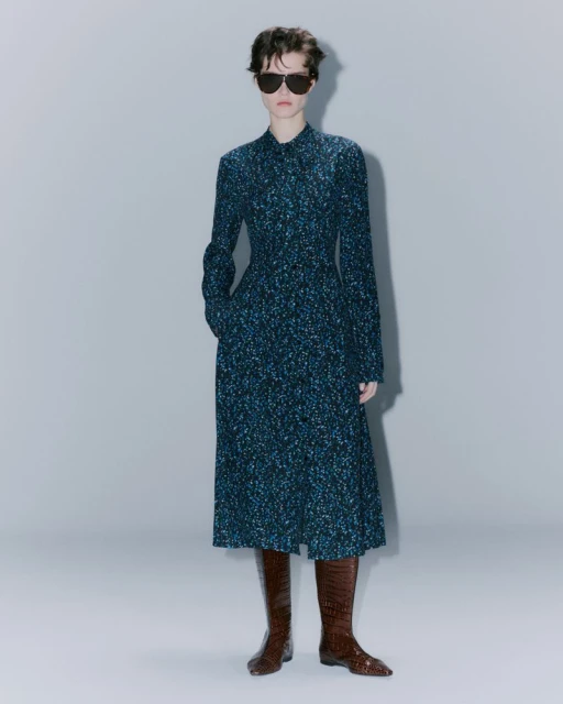 Full-length shot of a stylish woman in a patterned midi dress and brown leather boots with sunglasses against a neutral backdrop.