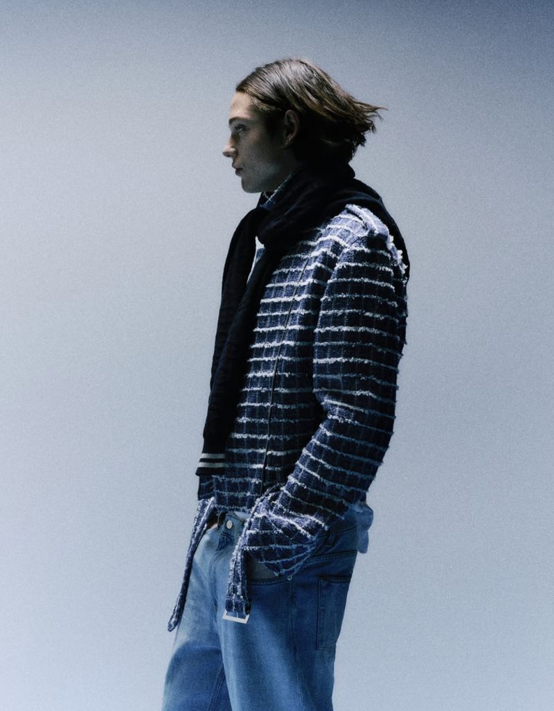 A young adult man in a dark jacket and jeans, standing in a studio setting with soft lighting, creating a fashion-forward and stylish look.