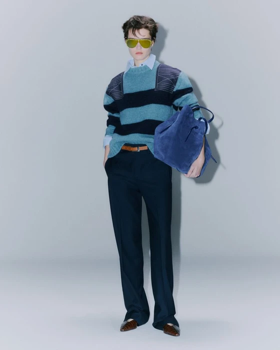Model wearing yellow sunglasses, striped sweater, and dark trousers holding a blue bag against a seamless light grey background.