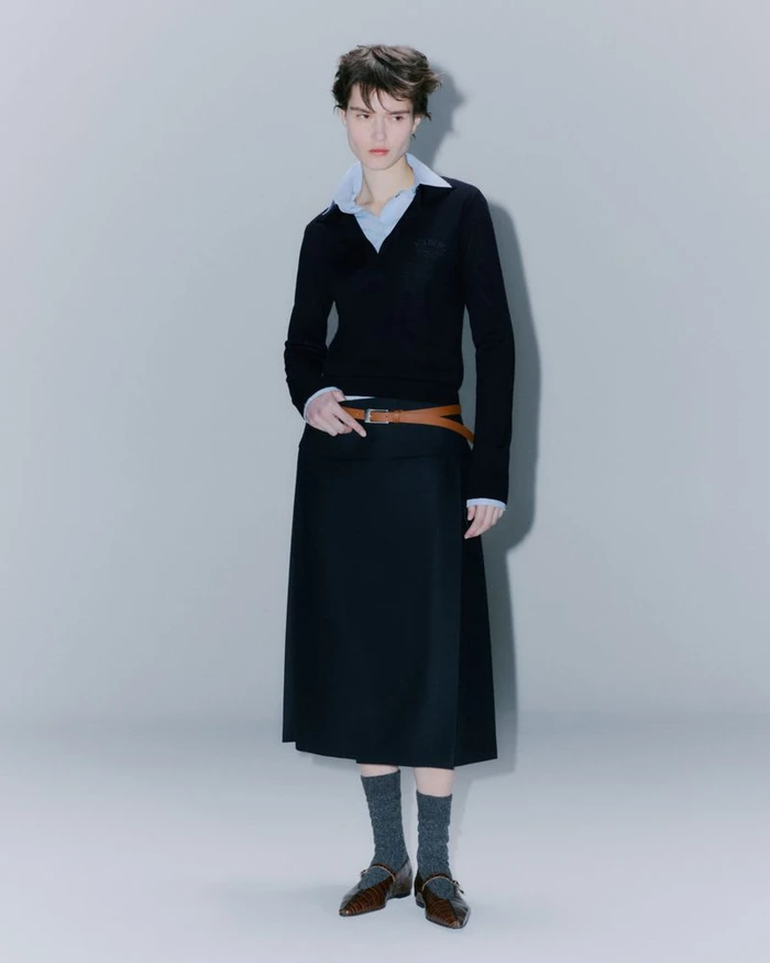 Model in black V-neck sweater and dark midi skirt with a brown belt against a plain grey background