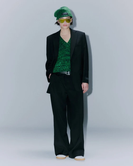 Model wearing a black blazer, green sweater, and green beanie against a grey seamless background.