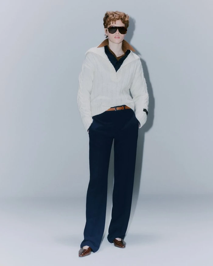 Model wearing a white cable-knit zip sweater, dark trousers, and sunglasses standing against a plain grey backdrop.