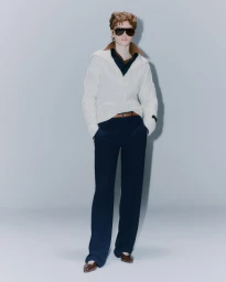 Model wearing a white cable-knit zip sweater, dark trousers, and sunglasses standing against a plain grey backdrop.