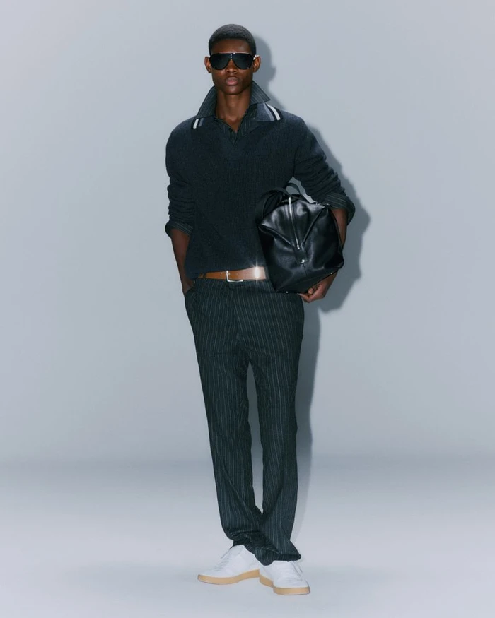 Model wearing dark sunglasses, dark polo sweater, and pinstripe trousers holding a black leather bag against a grey seamless background