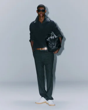 Model wearing dark sunglasses, dark polo sweater, and pinstripe trousers holding a black leather bag against a grey seamless background