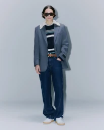 Model wearing dark sunglasses, grey blazer over striped sweater, blue jeans, and white sneakers against a grey backdrop