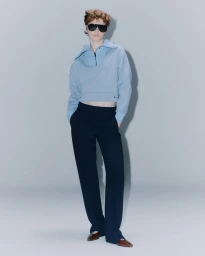 Model in dark sunglasses, light blue half-zip sweatshirt, and dark trousers standing against a pale grey studio background.