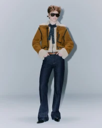 Model in dark sunglasses wearing a brown shearling jacket and dark denim jeans against a grey studio backdrop.