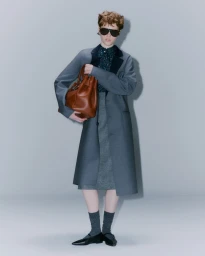 Model wearing a long grey coat, dark sunglasses, and holding a brown leather handbag against a plain grey seamless background.