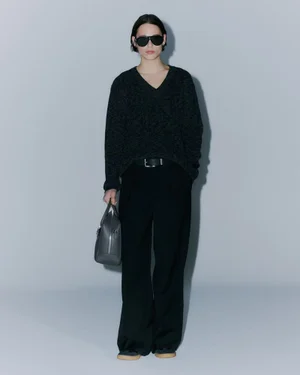 Model in black sunglasses, dark sweater, and trousers holding a grey handbag against a seamless grey studio background.