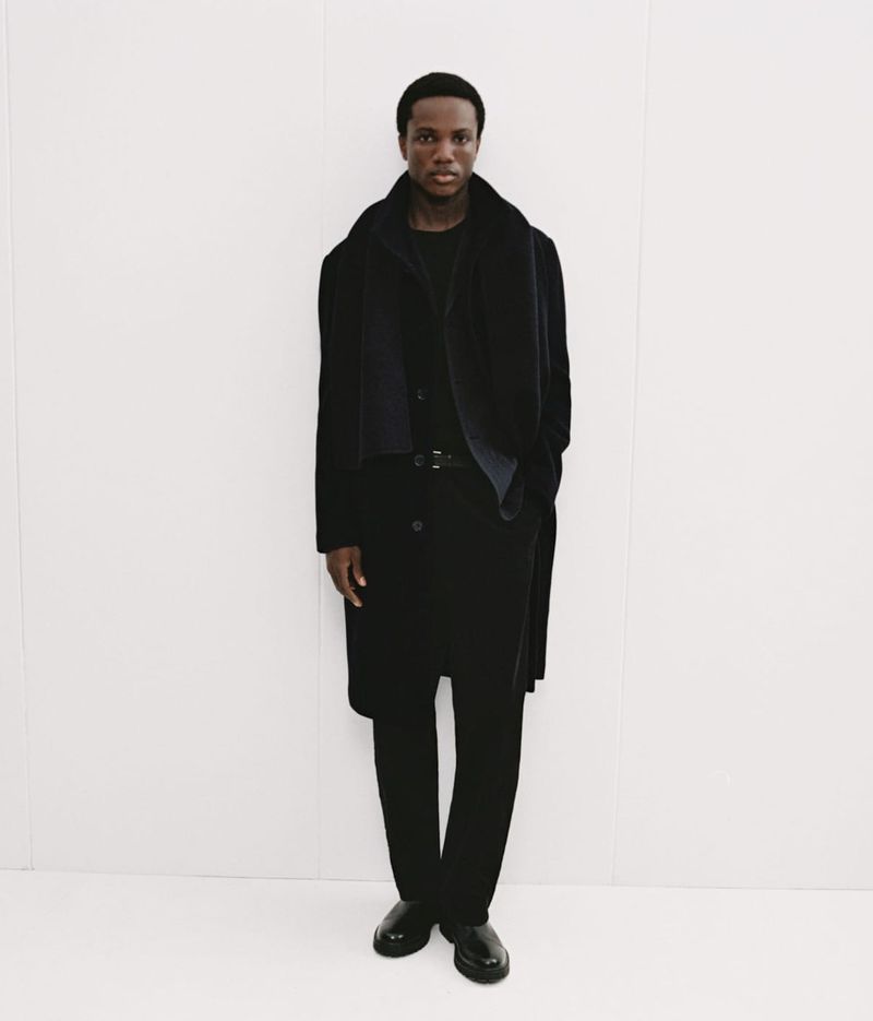 Black man wearing a dark coat and trousers posing against a bright white background for Mango Winter Wardrobe.