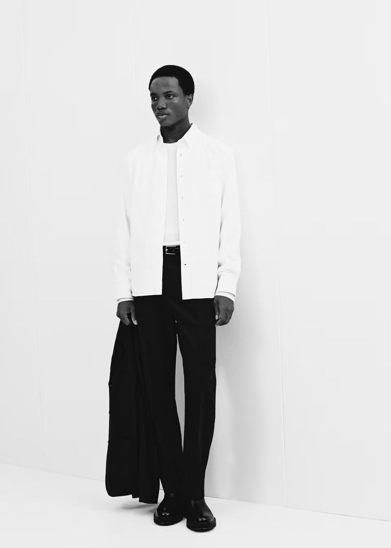 Black male model in white button-down shirt, black trousers, and black leather shoes posing in front of a white wall in a photo studio.