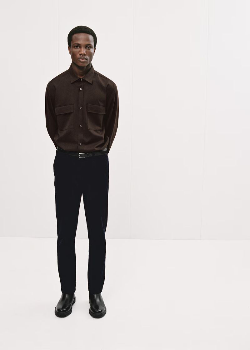 Man wearing a brown long-sleeve shirt and dark trousers standing against a plain white studio background.