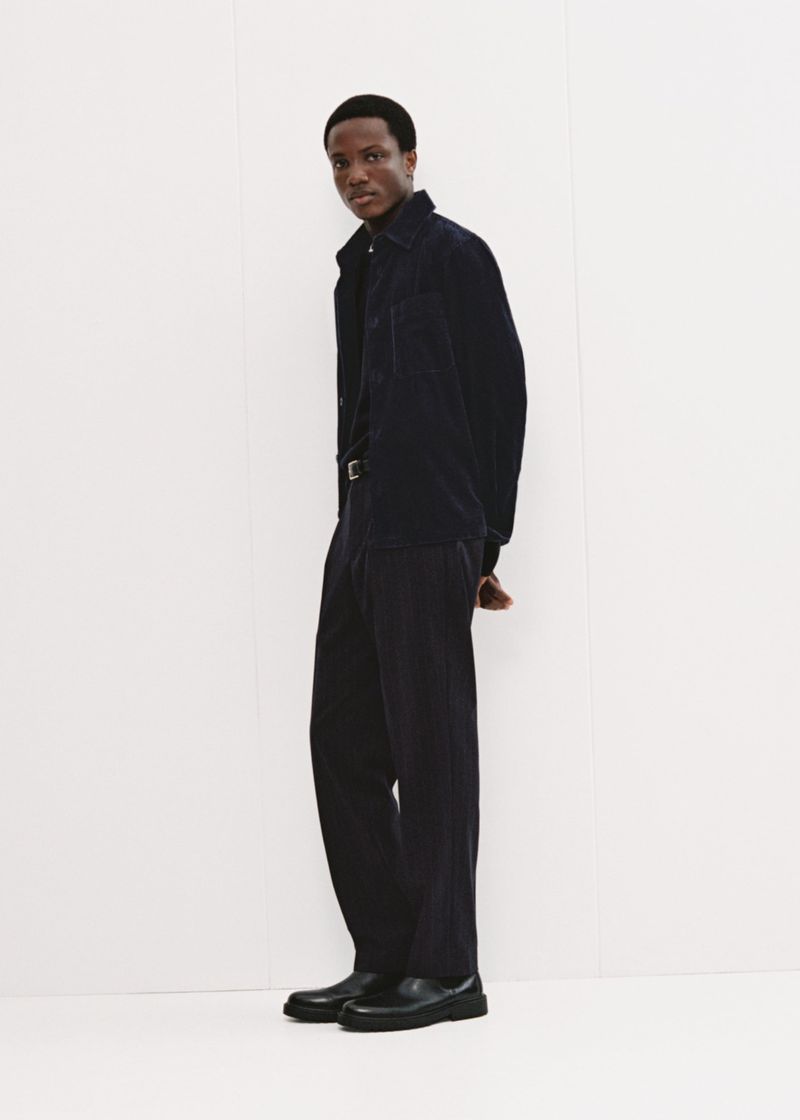 Full-length shot of an adult Black man wearing a dark blue corduroy shirt and pants with black shoes, standing against a white wall.