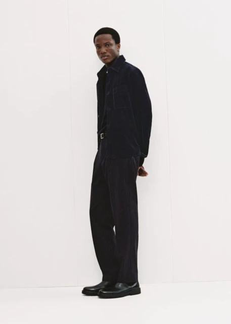 Full-length shot of an adult Black man wearing a dark blue corduroy shirt and pants with black shoes, standing against a white wall.