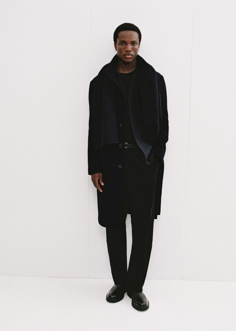 Model in dark coat and black scarf standing against a plain white background for Mango Winter Wardrobe.