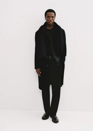 Model in dark coat and black scarf standing against a plain white background for Mango Winter Wardrobe.
