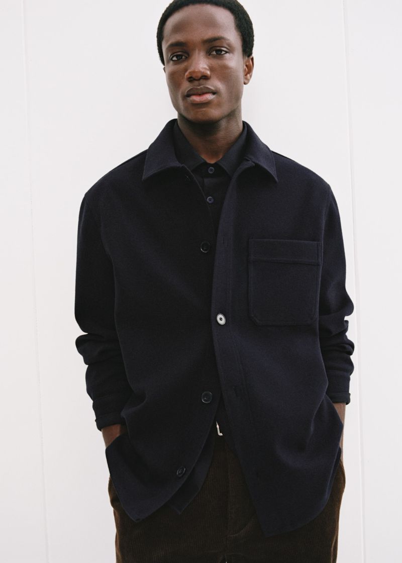 Dark-skinned male model poses in a navy button-down shirt and brown pants in a bright, neutral-toned photo studio, for a fashion editorial.