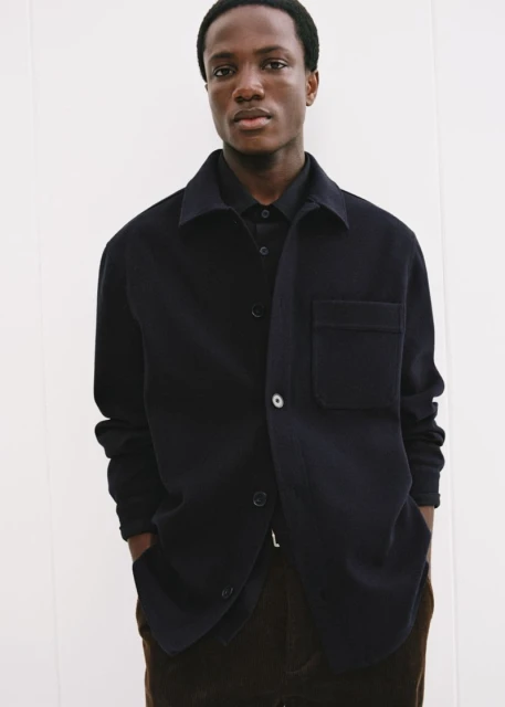 Dark-skinned male model poses in a navy button-down shirt and brown pants in a bright, neutral-toned photo studio, for a fashion editorial.