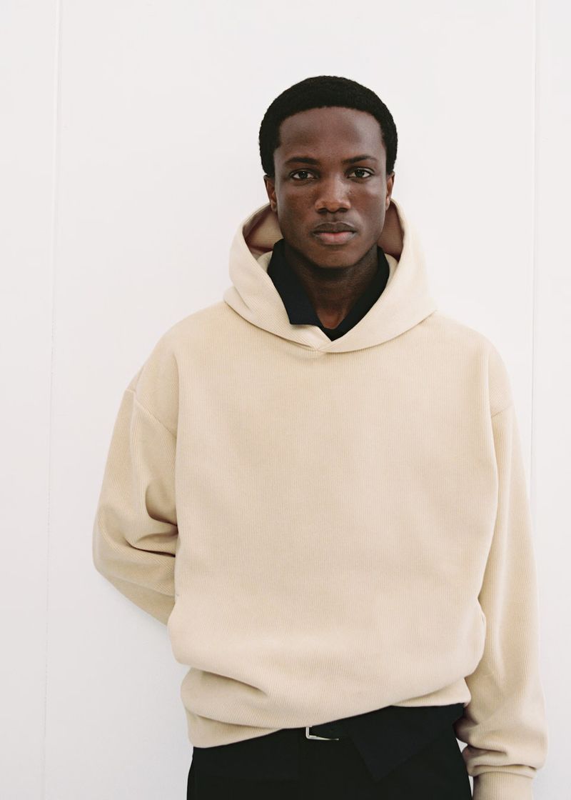 Man wearing a beige hoodie stands against a white wall, showcasing fashion apparel in a simple indoor studio setting with soft lighting.
