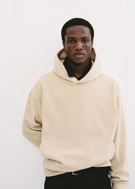 Man wearing a beige hoodie stands against a white wall, showcasing fashion apparel in a simple indoor studio setting with soft lighting.