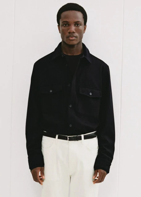 Man in black button-down shirt and white pants poses in a studio with neutral background. Fashion shot, portrait orientation.