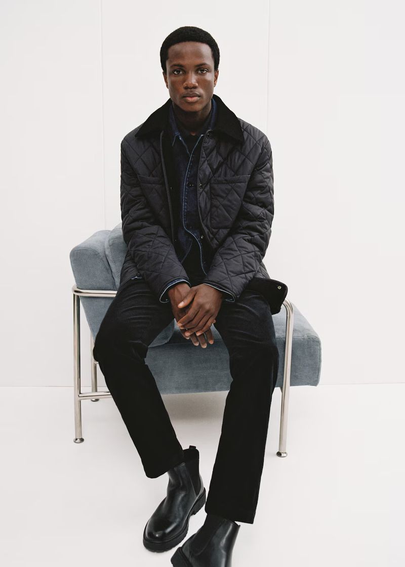 Model Mumin Jangani seated in a blue armchair wearing a black quilted jacket and dark trousers for Mango Winter Wardrobe.