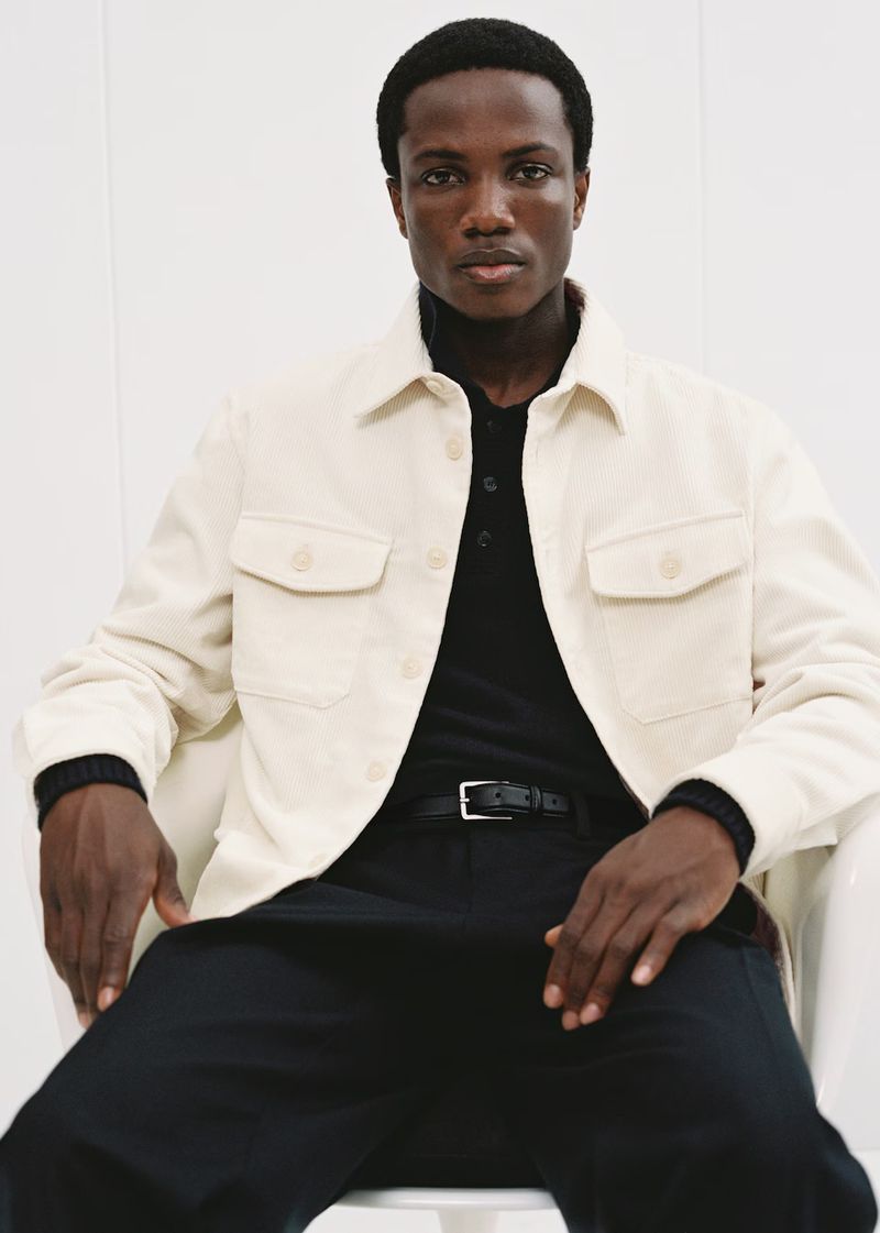 A stylish dark-skinned man is sitting in a bright studio wearing a beige chore jacket over a black polo shirt and dark pants.