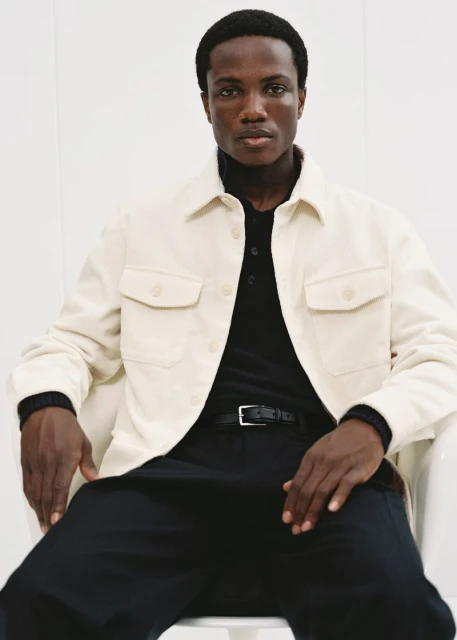 A stylish dark-skinned man is sitting in a bright studio wearing a beige chore jacket over a black polo shirt and dark pants.