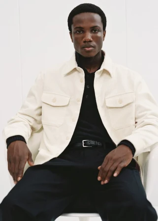 A stylish dark-skinned man is sitting in a bright studio wearing a beige chore jacket over a black polo shirt and dark pants.