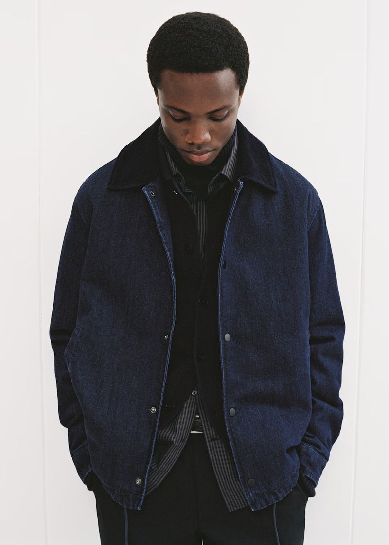 A stylish African-American man in a dark denim jacket and black turtleneck sweater poses against a plain white wall in a fashion shot.