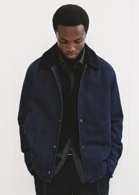 A stylish African-American man in a dark denim jacket and black turtleneck sweater poses against a plain white wall in a fashion shot.