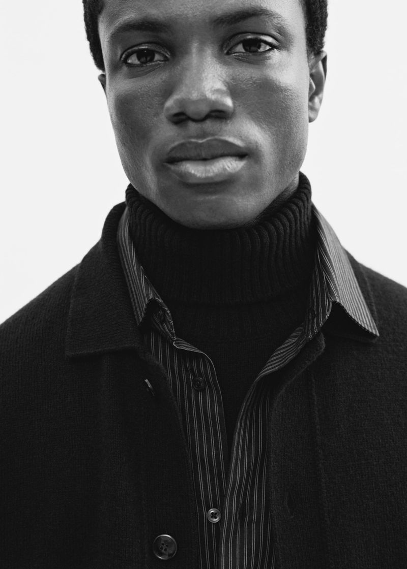 Portrait of a Black man in a black turtleneck and button-down shirt layered under a dark coat, set against a bright white backdrop.