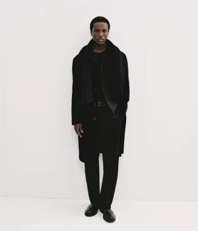 Black man wearing a dark coat and trousers posing against a bright white background for Mango Winter Wardrobe.
