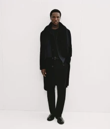 Black man wearing a dark coat and trousers posing against a bright white background for Mango Winter Wardrobe.
