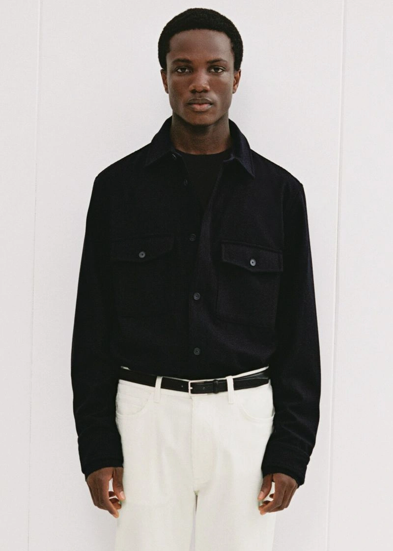 Black man wearing a dark long-sleeve button-up shirt and white trousers against a plain white studio background.