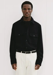 Black man wearing a dark long-sleeve button-up shirt and white trousers against a plain white studio background.