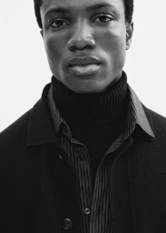 Close-up portrait of a Black man wearing a black turtleneck and button-up shirt against a plain light background