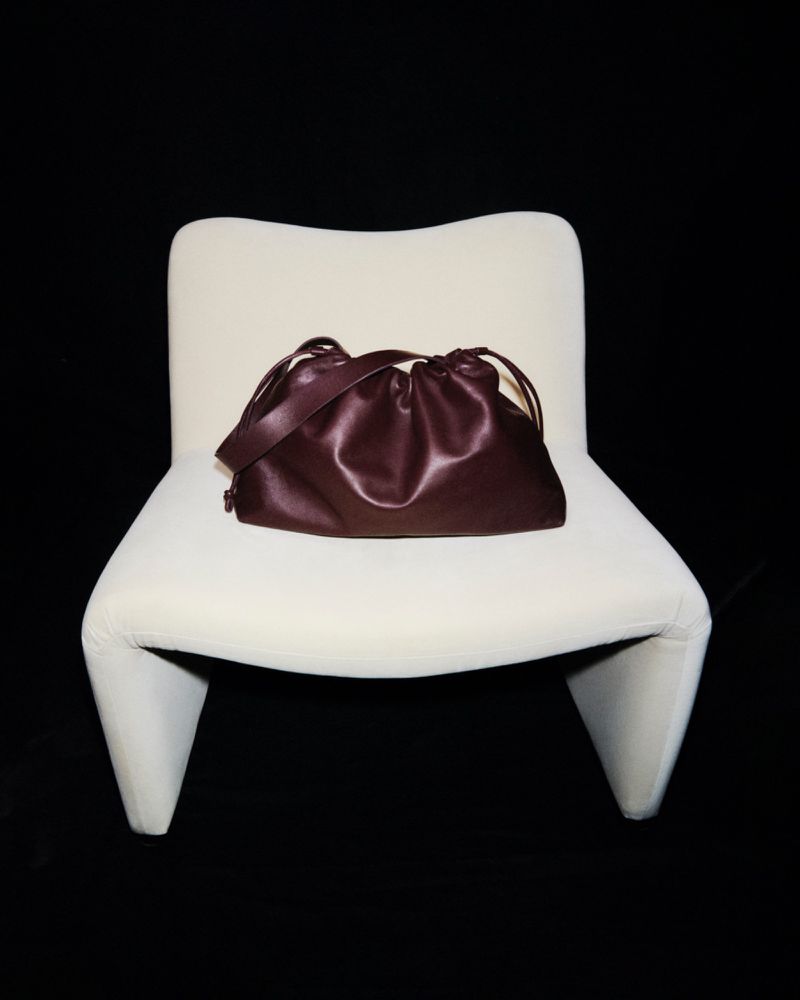 Burgundy satchel handbag with a drawstring closure and a shoulder strap sitting on a white modern chair against a black backdrop.