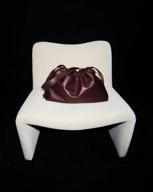 Burgundy satchel handbag with a drawstring closure and a shoulder strap sitting on a white modern chair against a black backdrop.
