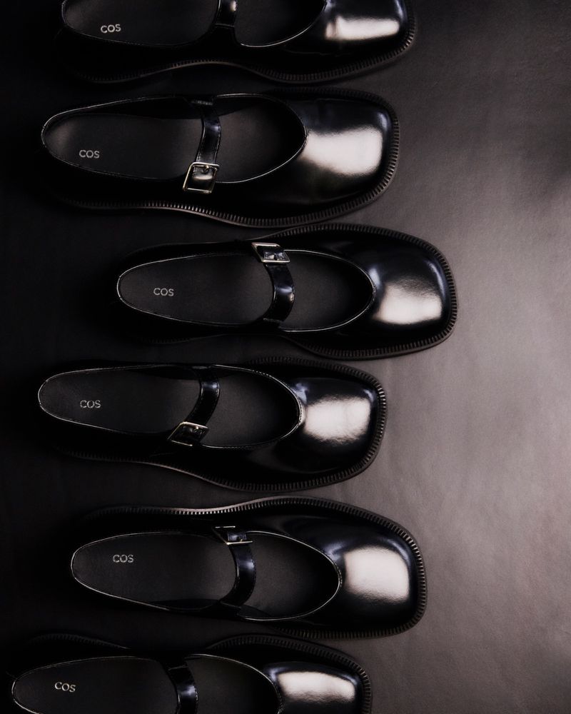 Overhead shot of multiple pairs of black COS brand leather Mary Jane shoes, arranged in a row on a dark surface for a fashion product display.