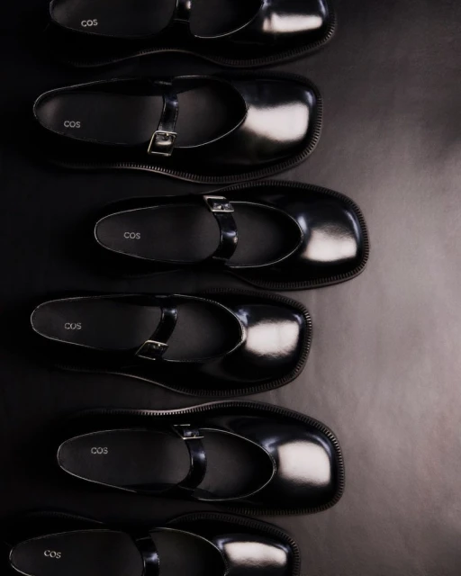 Overhead shot of multiple pairs of black COS brand leather Mary Jane shoes, arranged in a row on a dark surface for a fashion product display.