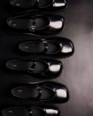 Overhead shot of multiple pairs of black COS brand leather Mary Jane shoes, arranged in a row on a dark surface for a fashion product display.